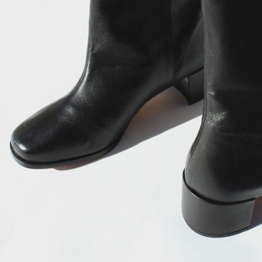 About Arianne black leather pull on boot with almond shaped toe, block heel, and v- shaped dip detail at top of shaft
