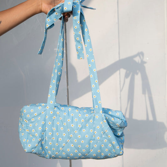 Blue daisy quilted duffle bag with shoulder straps that tie into bows. Zipper closure. Available at Ease Toronto