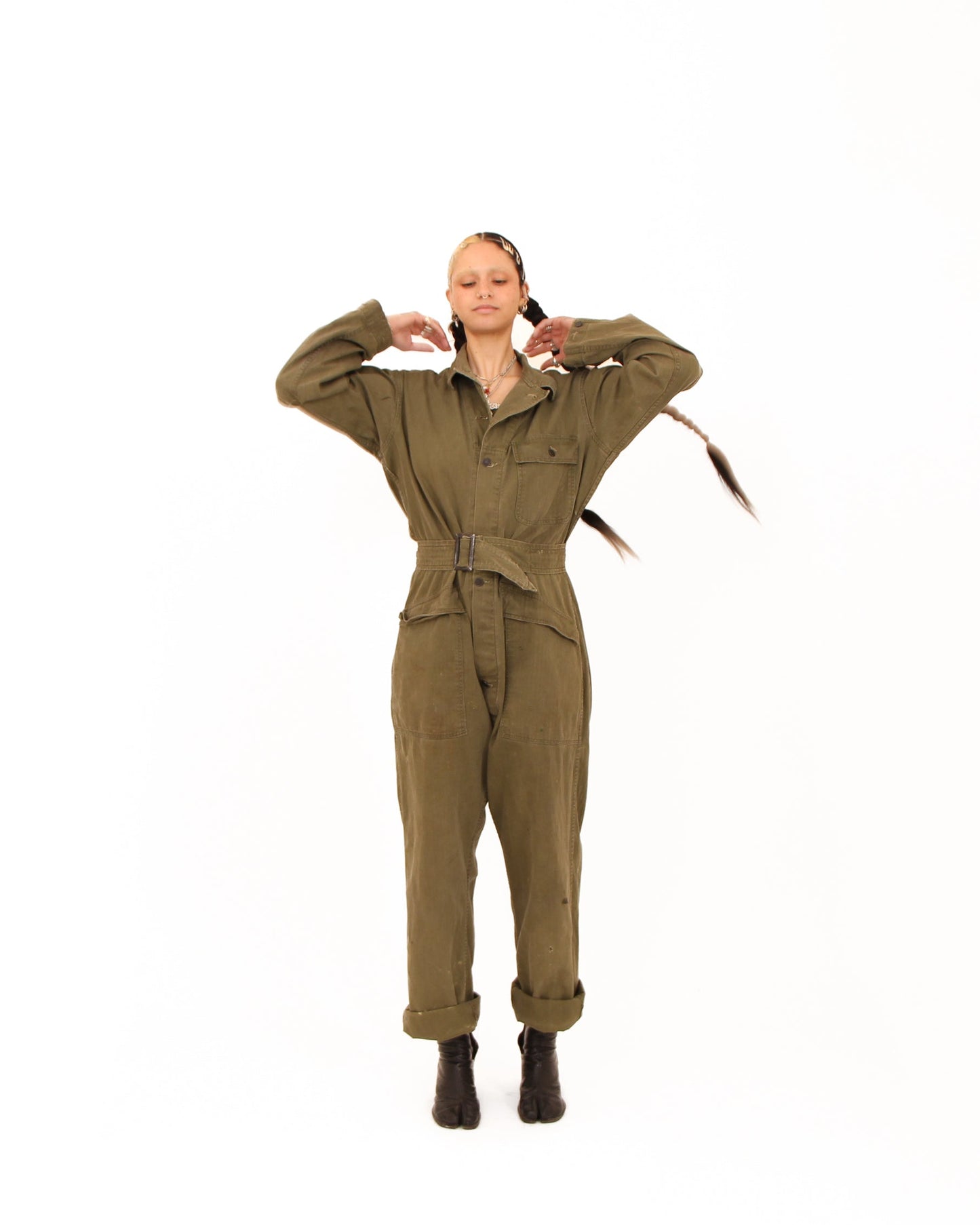 A woman modeling an olive green military-style jumpsuit with a matching waist tie, standing against a solid white background.