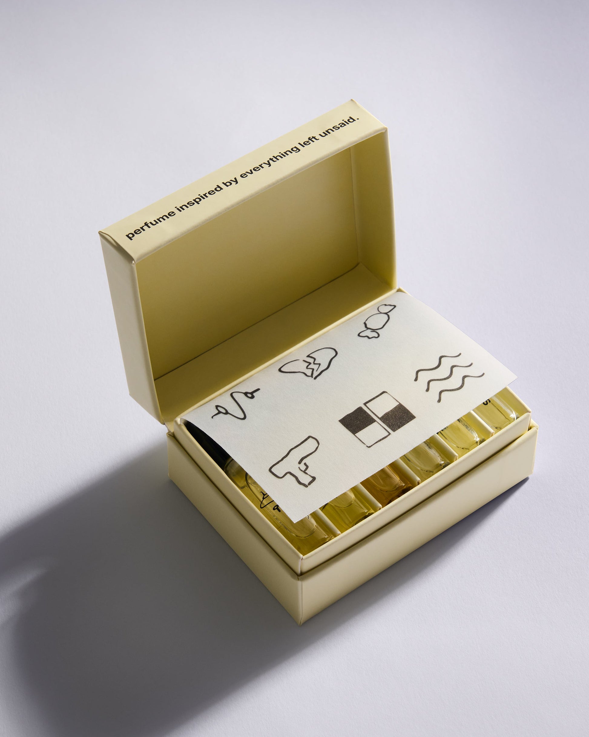 An open pale yellow perfume box on a light gray background, showing a folded cream-colored card with minimalist black line drawings and a row of gold-toned rectangular items inside.