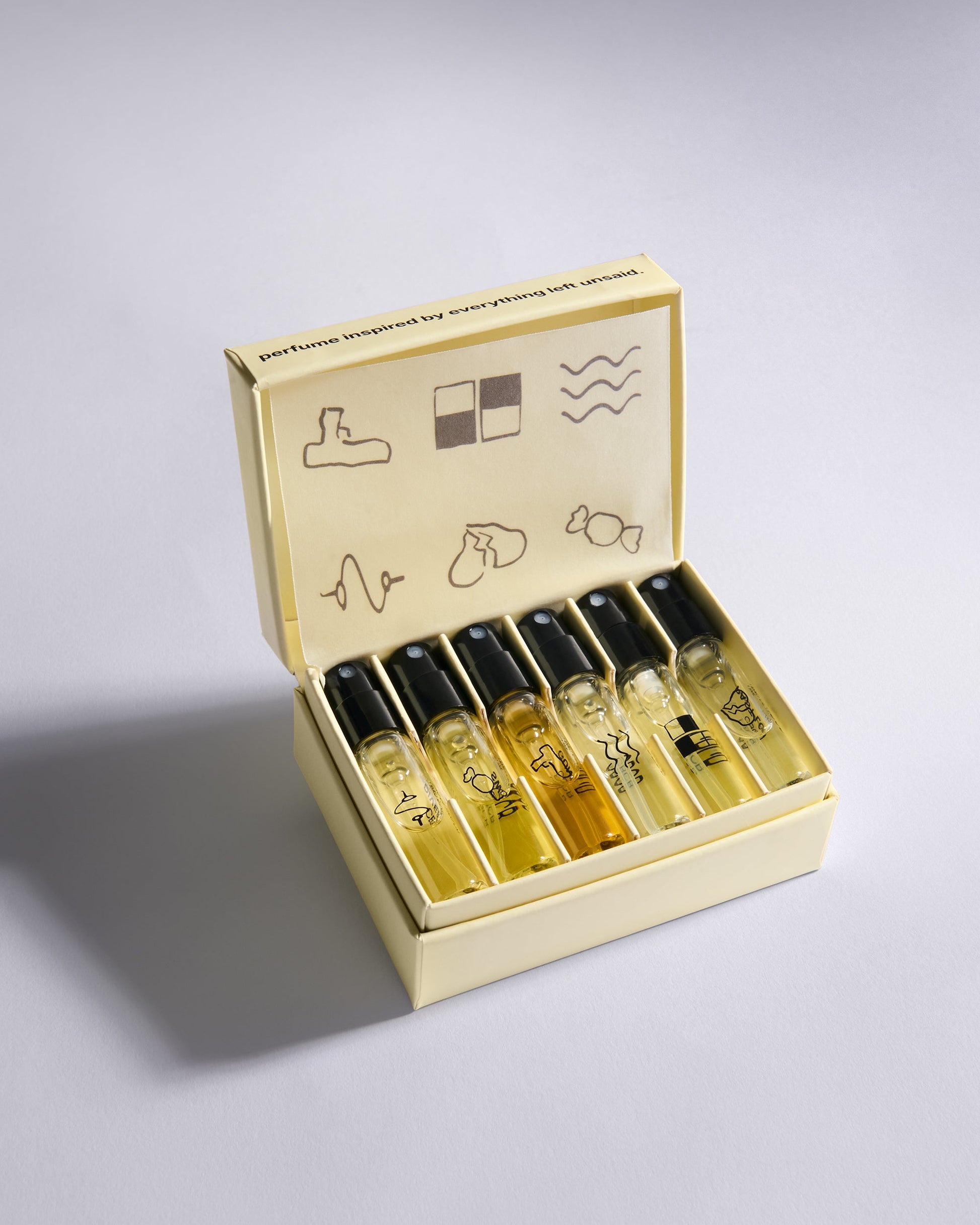 A set of six perfume sample vials with black caps arranged inside an open cream-colored presentation box on a light gray background.