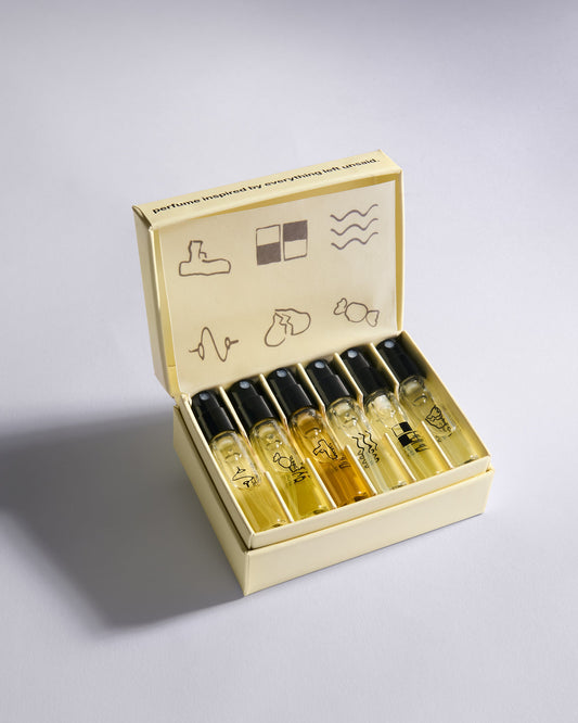 A set of six perfume sample vials with black caps arranged inside an open cream-colored presentation box on a light gray background.