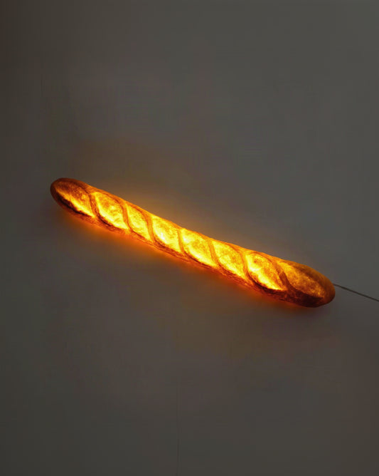 An illuminated bread-shaped light fixture shaped like a baguette, glowing with a warm orange-yellow light against a dark gray background.