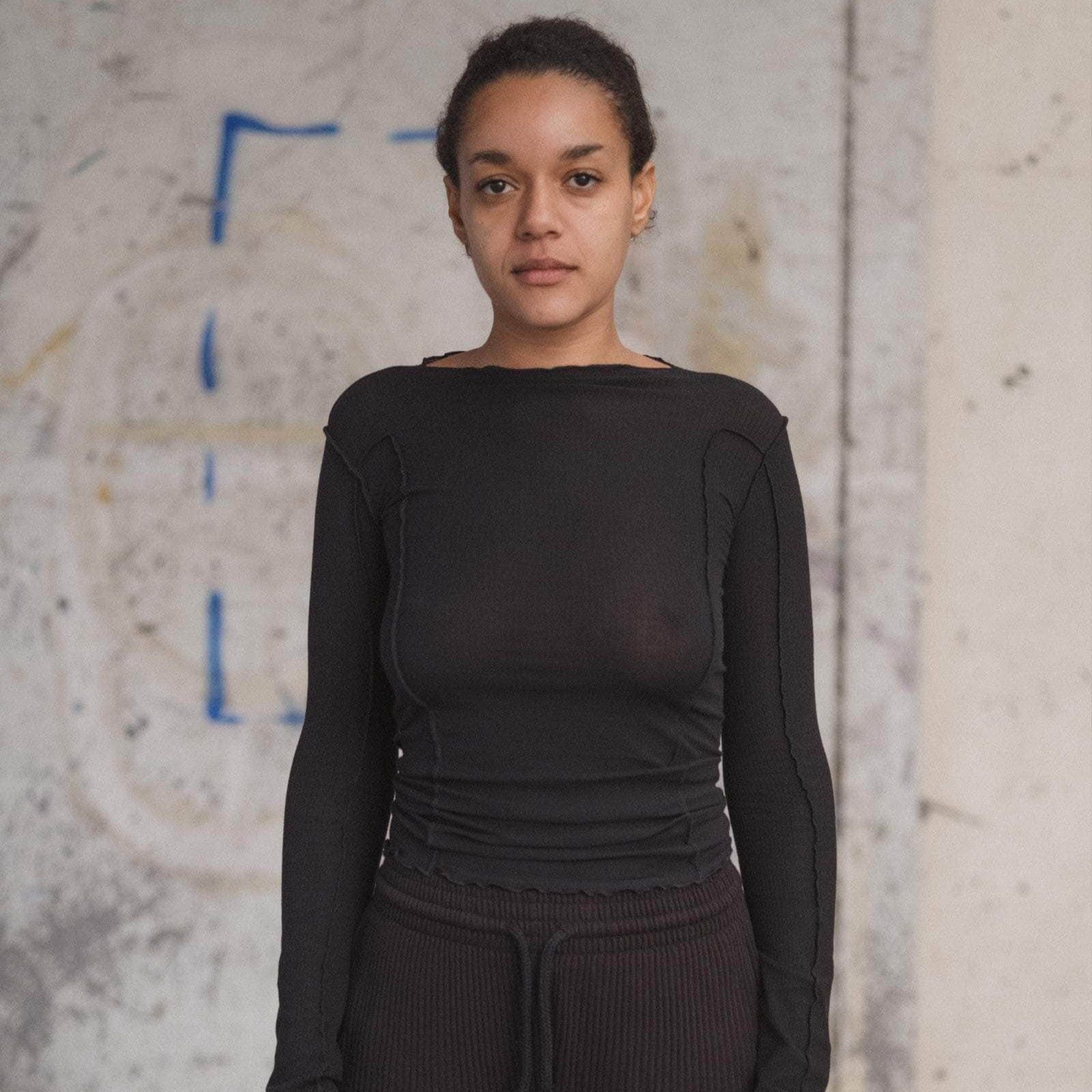 Person wearing a black long-sleeve top and dark pants against a textured wall.