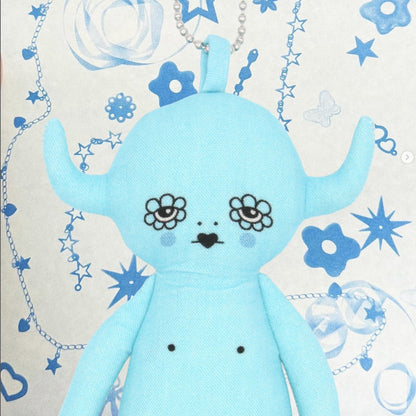 Blue plush toy devil charm with floral designs on a decorative background with stars and flowers.