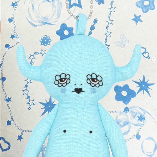 Blue plush toy devil charm with floral designs on a decorative background with stars and flowers.