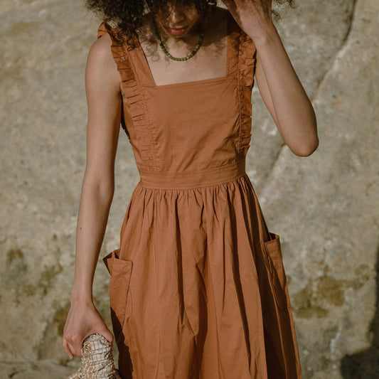 Cinamon coloured apron dress with pockets, defined waist band, large straps at back to tie as bow detail, ruffled shoulder straps, and square neckline