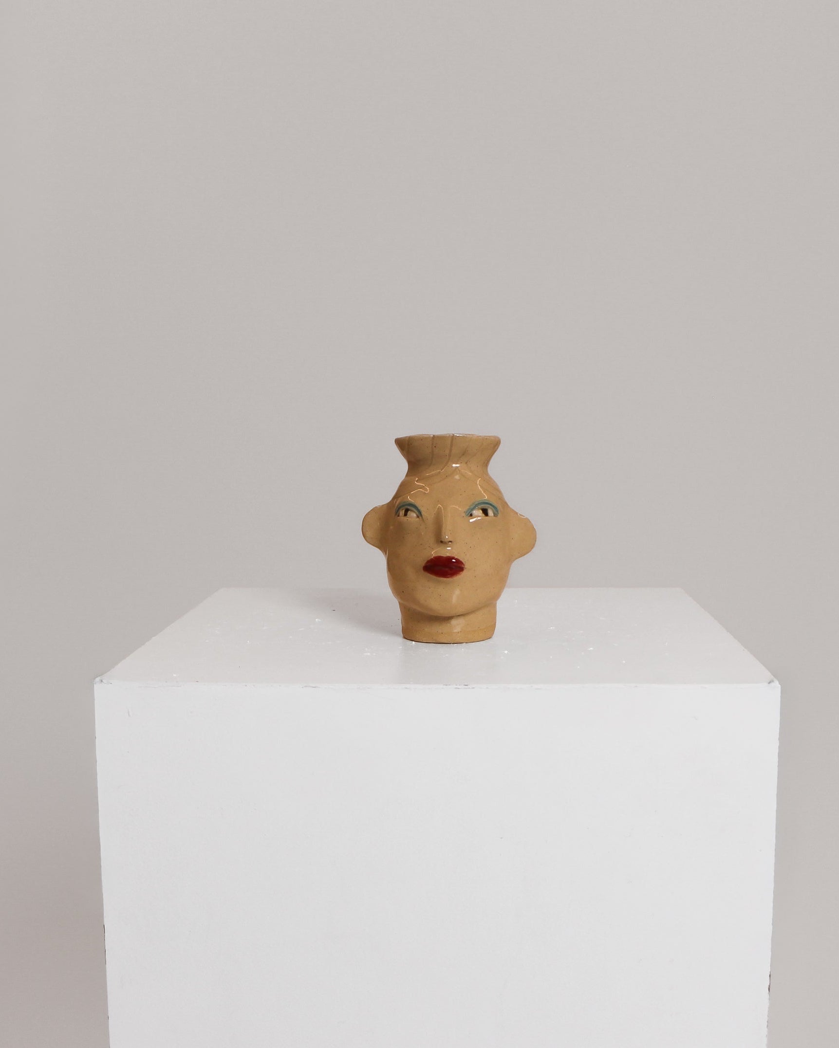 A small sculptural ceramic vase shaped like a stylized female head with red lips and painted eyes, displayed on a white pedestal against a light gray background.