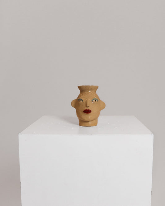A small sculptural ceramic vase shaped like a stylized female head with red lips and painted eyes, displayed on a white pedestal against a light gray background.