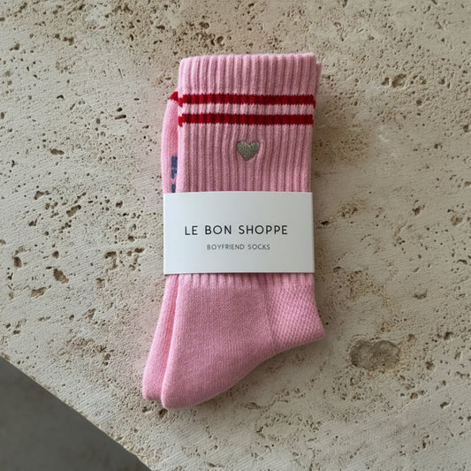 Pink socks with a heart design and red stripes on a beige stone surface, labeled 'Le Bon Shoppe'.