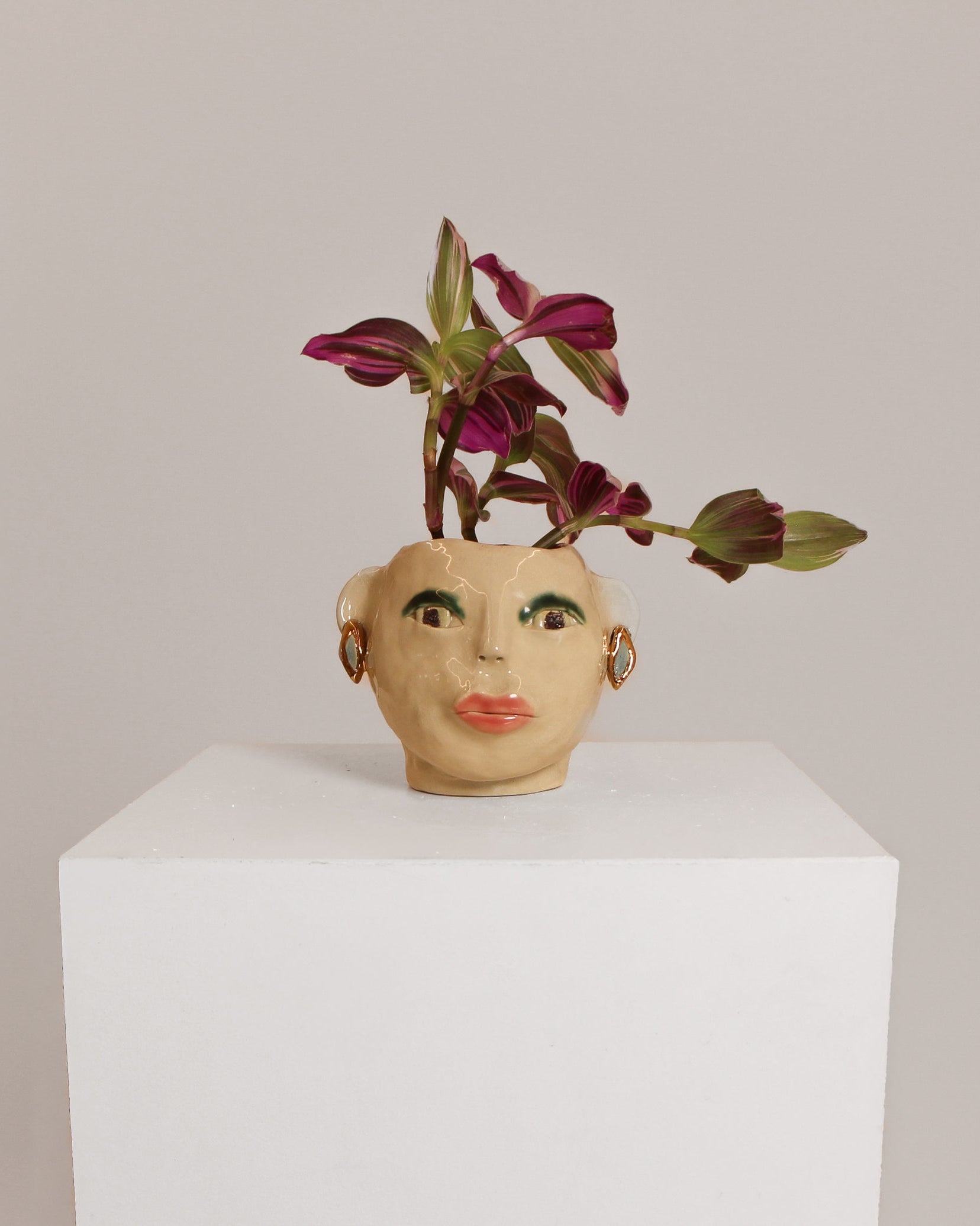 A ceramic face-shaped planter with painted facial features, holding a small purple and green trailing plant, displayed on a white pedestal against a light gray background.