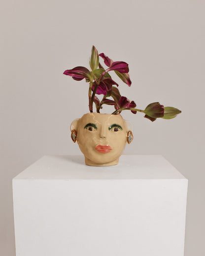 A ceramic face-shaped planter with painted facial features, holding a small purple and green trailing plant, displayed on a white pedestal against a light gray background.