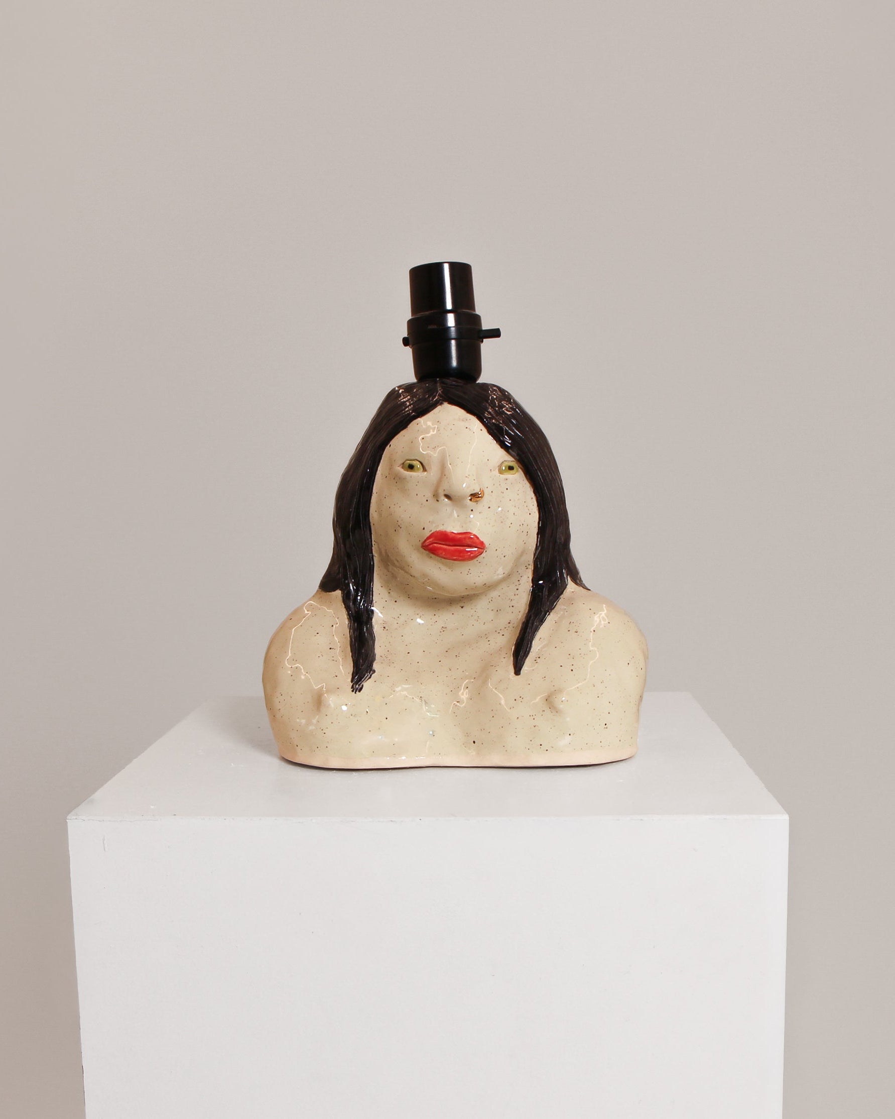 A ceramic sculpture of a woman's bust with dark hair, red lips, and a small black top hat, displayed on a white pedestal against a light gray background.