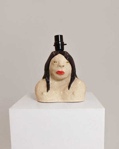 A ceramic sculpture of a woman's bust with dark hair, red lips, and a small black top hat, displayed on a white pedestal against a light gray background.