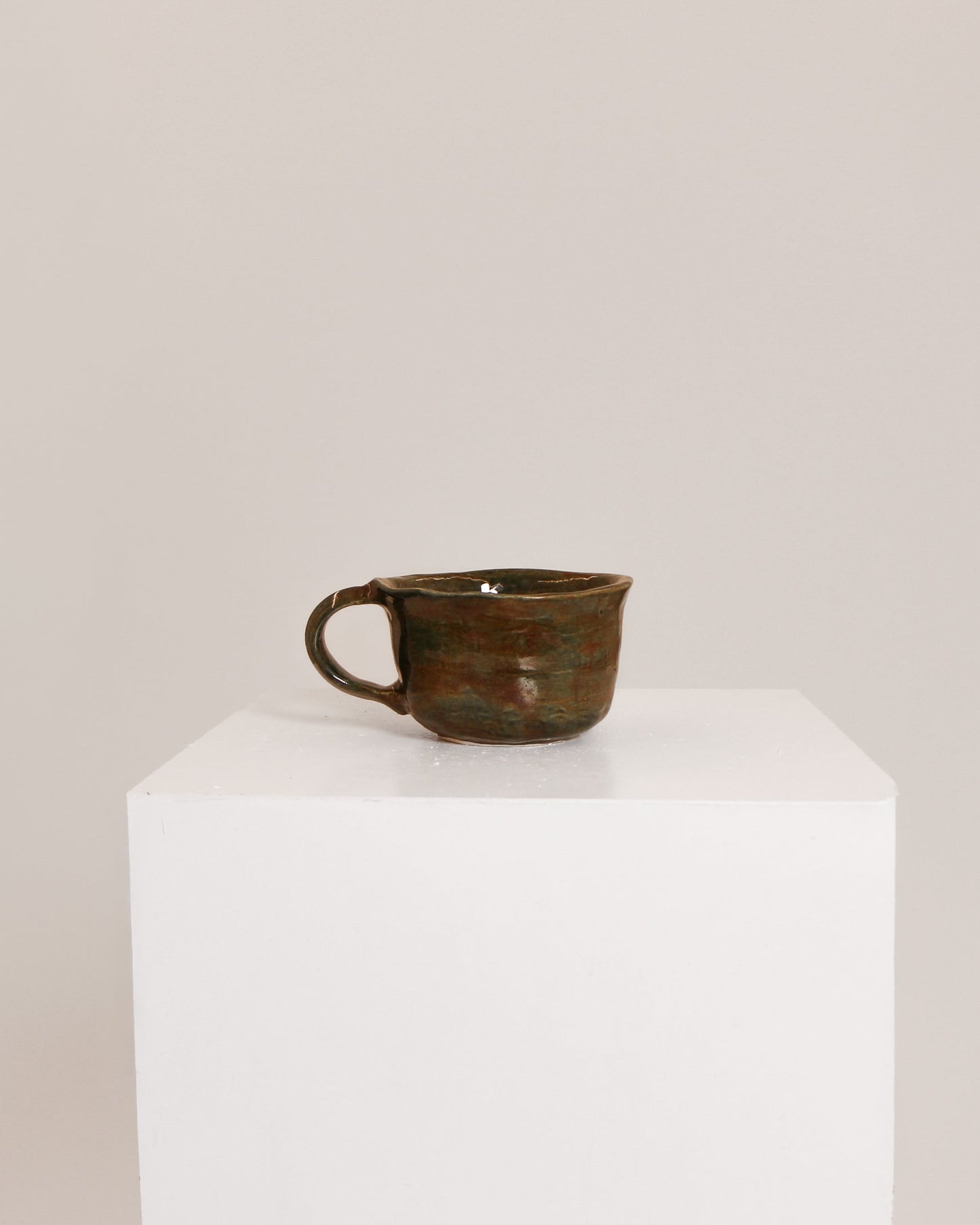 A small rustic ceramic cup with a dark brown and green glaze sits on a white display pedestal against a plain light beige background.