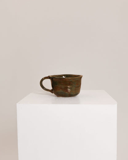 A small rustic ceramic cup with a dark brown and green glaze sits on a white display pedestal against a plain light beige background.