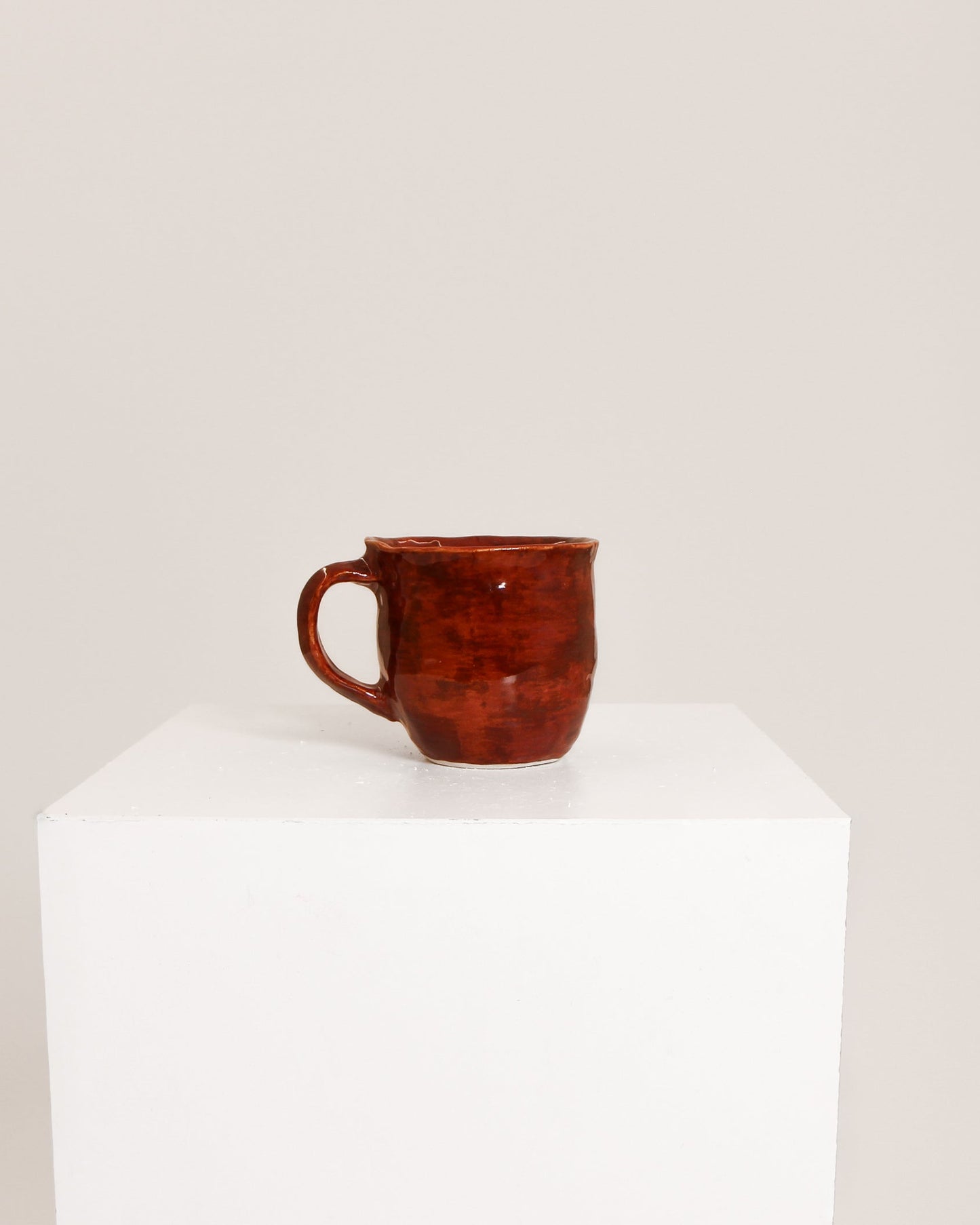 A small rustic ceramic mug with a reddish-brown glaze sits on a white display pedestal against a plain light beige background.