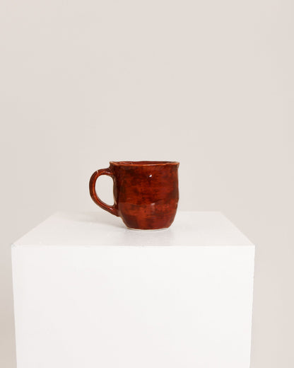 A small rustic ceramic mug with a reddish-brown glaze sits on a white display pedestal against a plain light beige background.