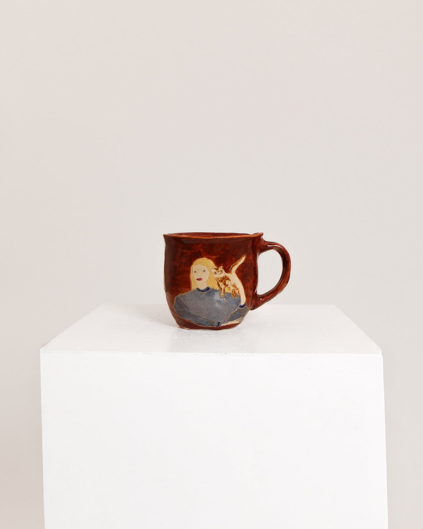 A small brown ceramic mug with a hand-painted illustration of a blonde woman holding a baby, displayed on a white pedestal against a light gray background.