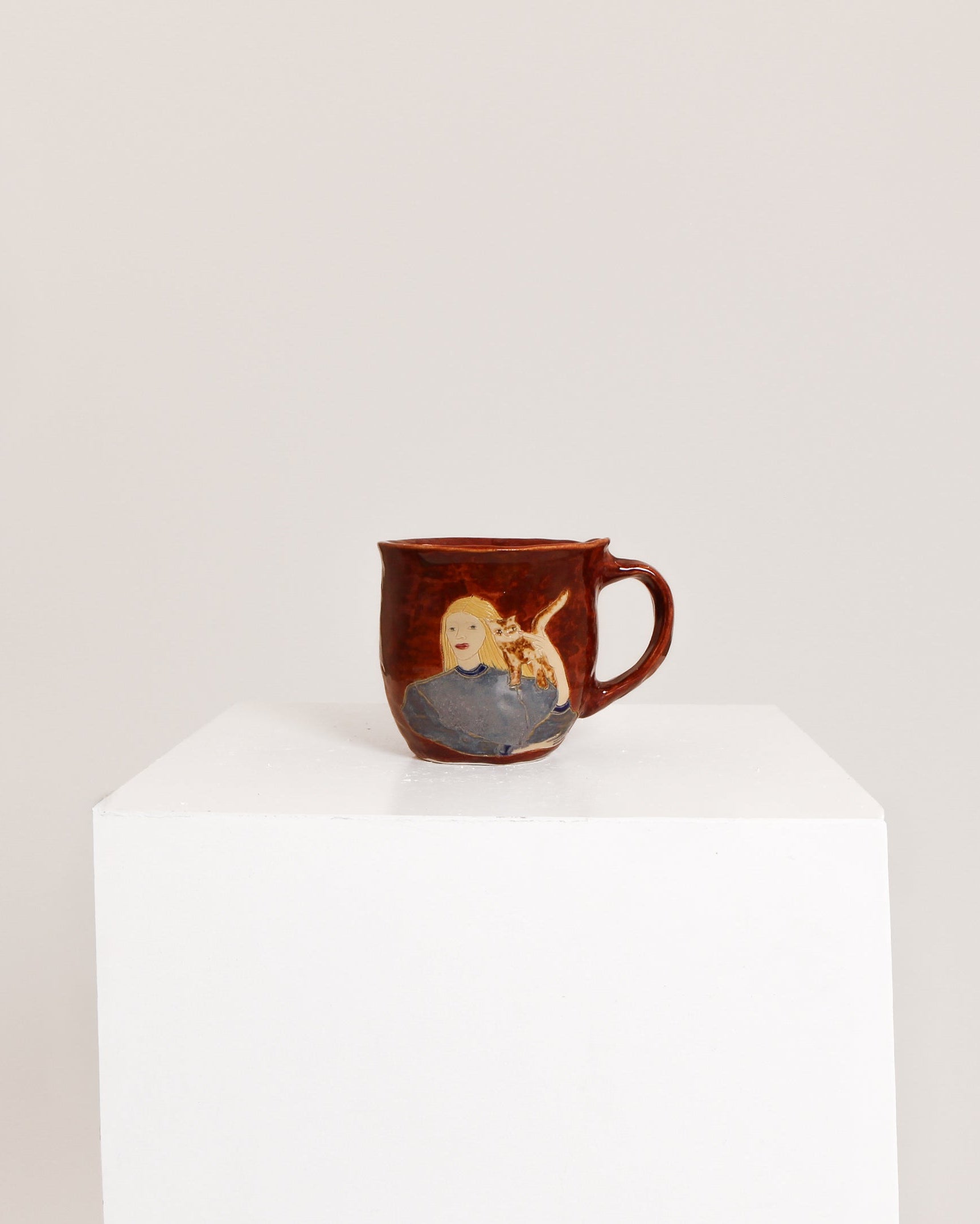 A small brown ceramic mug with a hand-painted illustration of a blonde woman holding a baby, displayed on a white pedestal against a light gray background.