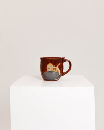 A small brown ceramic mug with a hand-painted illustration of a blonde woman holding a baby, displayed on a white pedestal against a light gray background.