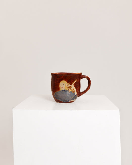 A small brown ceramic mug with a hand-painted illustration of a blonde woman holding a baby, displayed on a white pedestal against a light gray background.