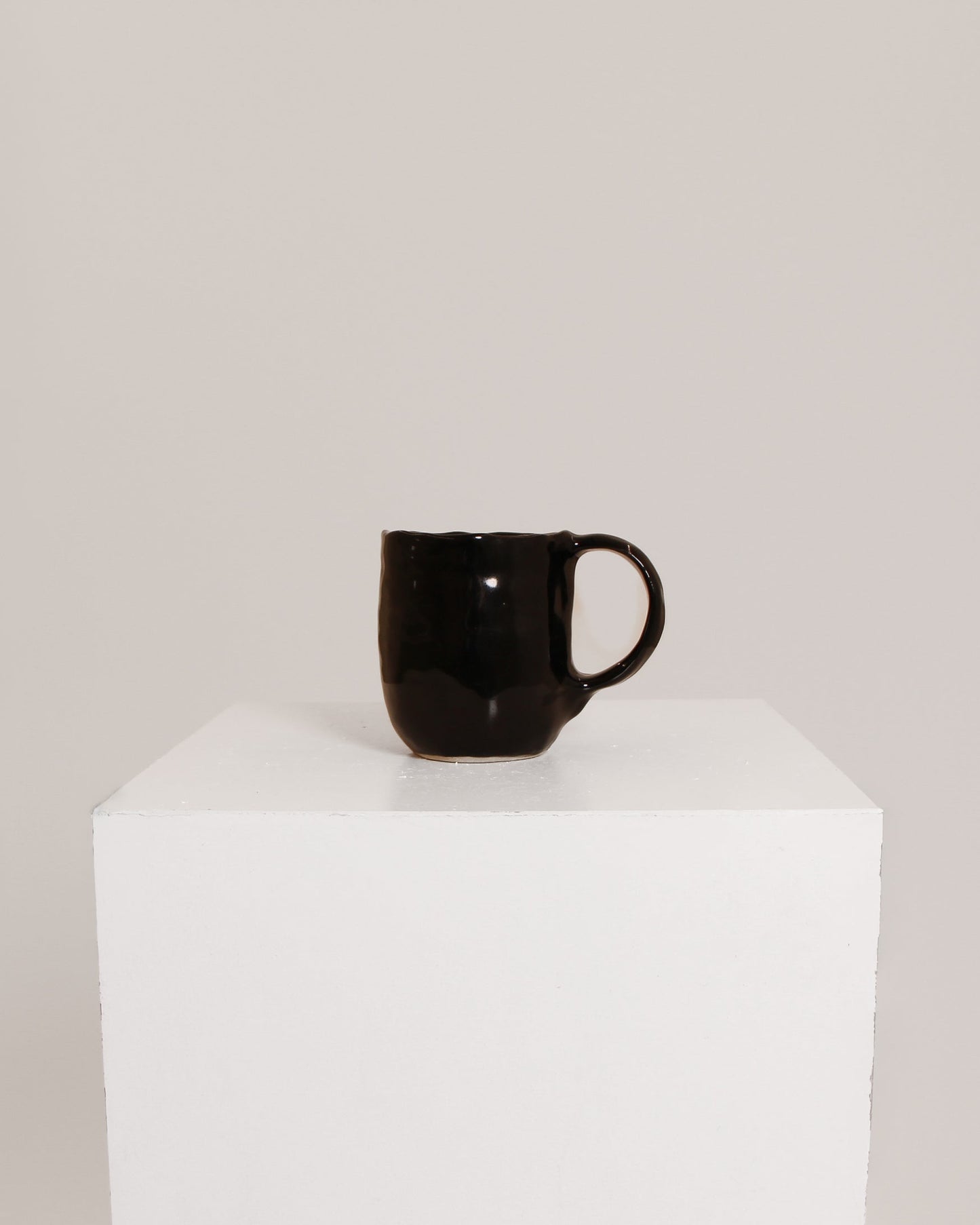 A glossy black ceramic mug with a curved handle, displayed on a white cube pedestal against a light gray background.