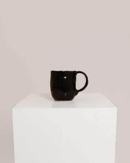 A glossy black ceramic mug with a curved handle, displayed on a white cube pedestal against a light gray background.