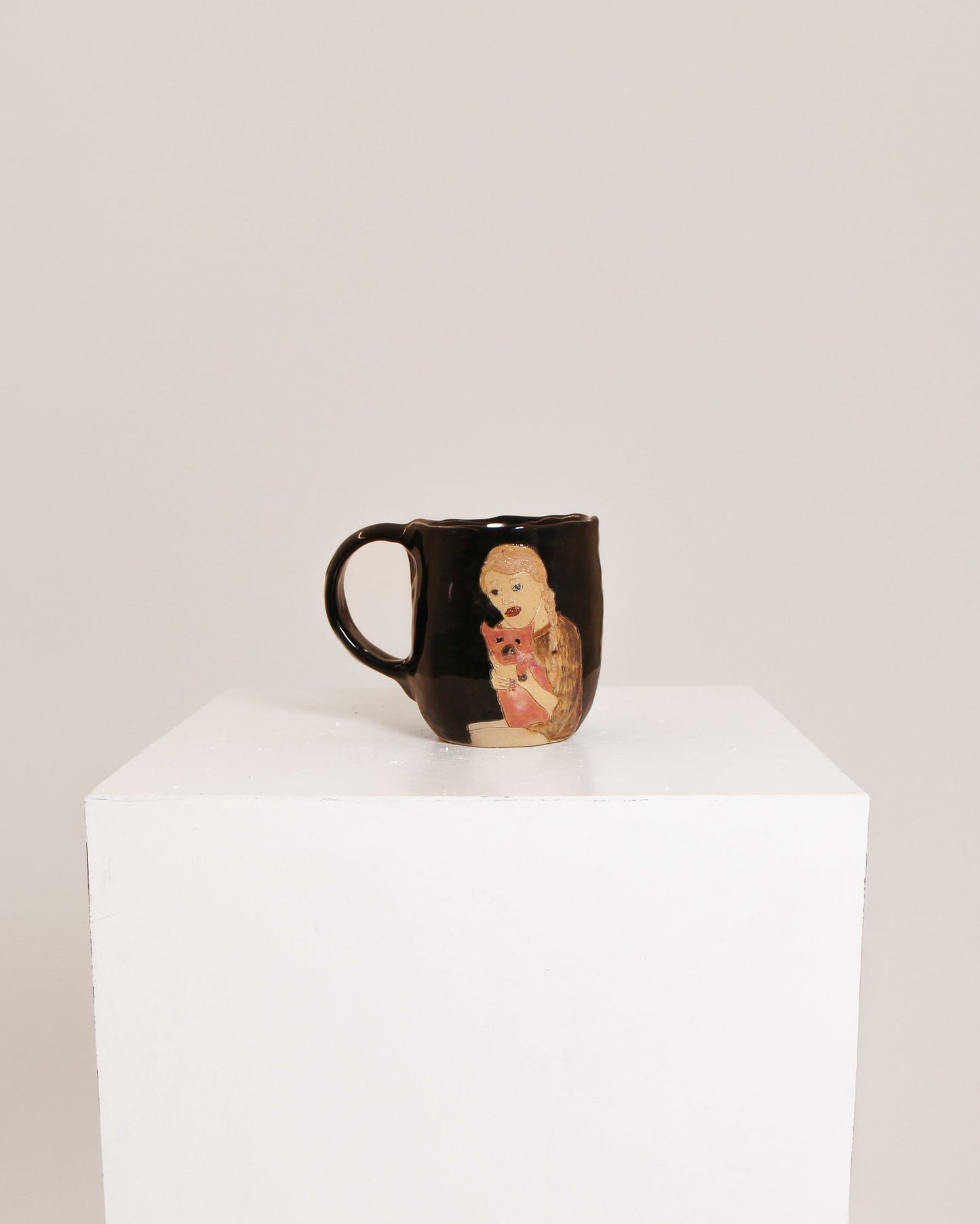 A glossy black ceramic mug featuring a hand-painted illustration of a baby doll, displayed on a white cube pedestal against a light gray background.