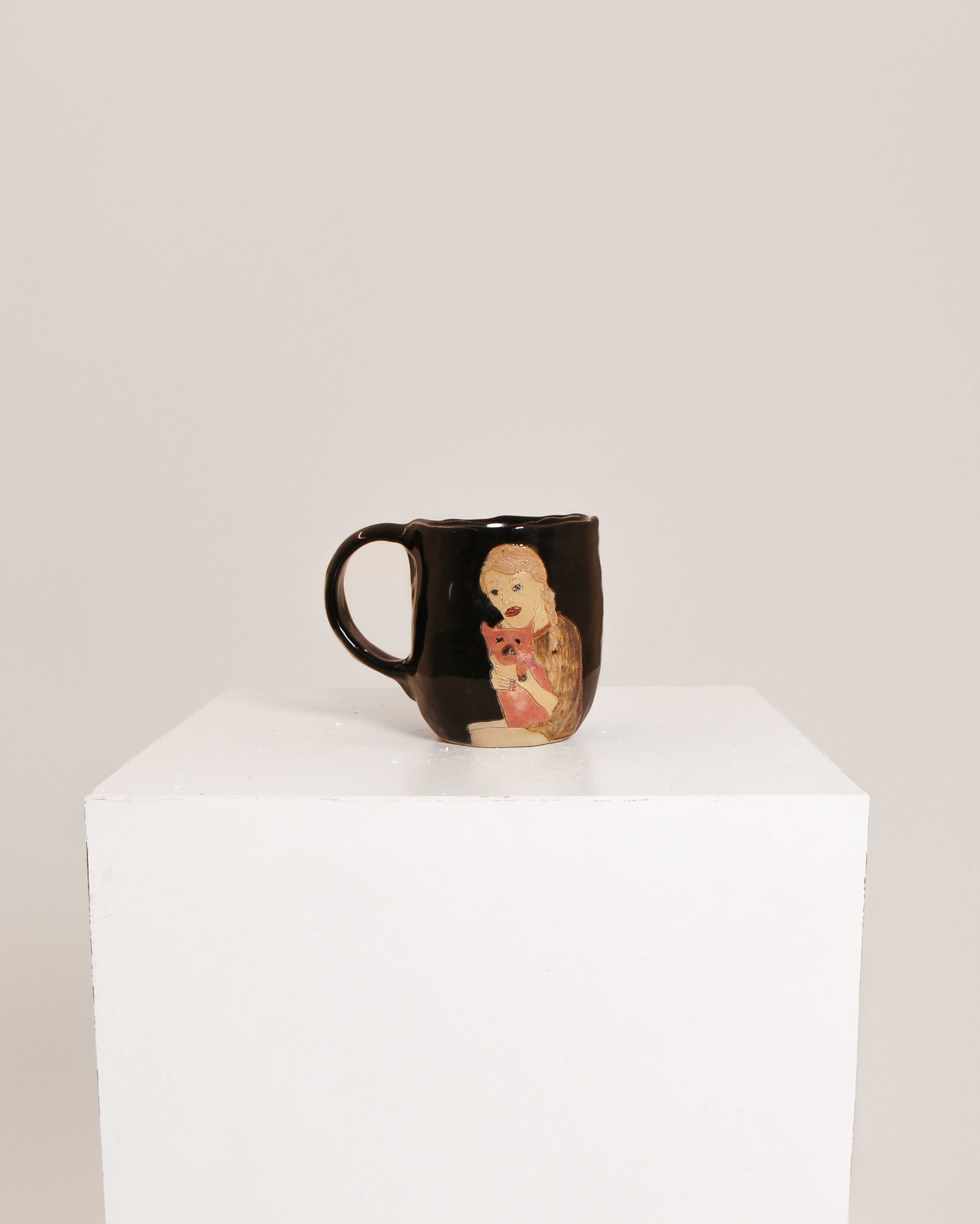 A glossy black ceramic mug featuring a hand-painted illustration of a baby doll, displayed on a white cube pedestal against a light gray background.