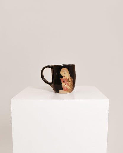 A glossy black ceramic mug featuring a hand-painted illustration of a baby doll, displayed on a white cube pedestal against a light gray background.