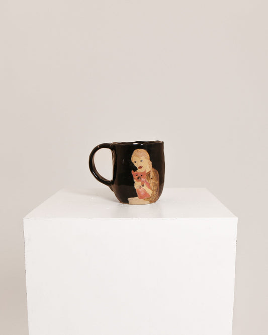 A glossy black ceramic mug featuring a hand-painted illustration of a baby doll, displayed on a white cube pedestal against a light gray background.