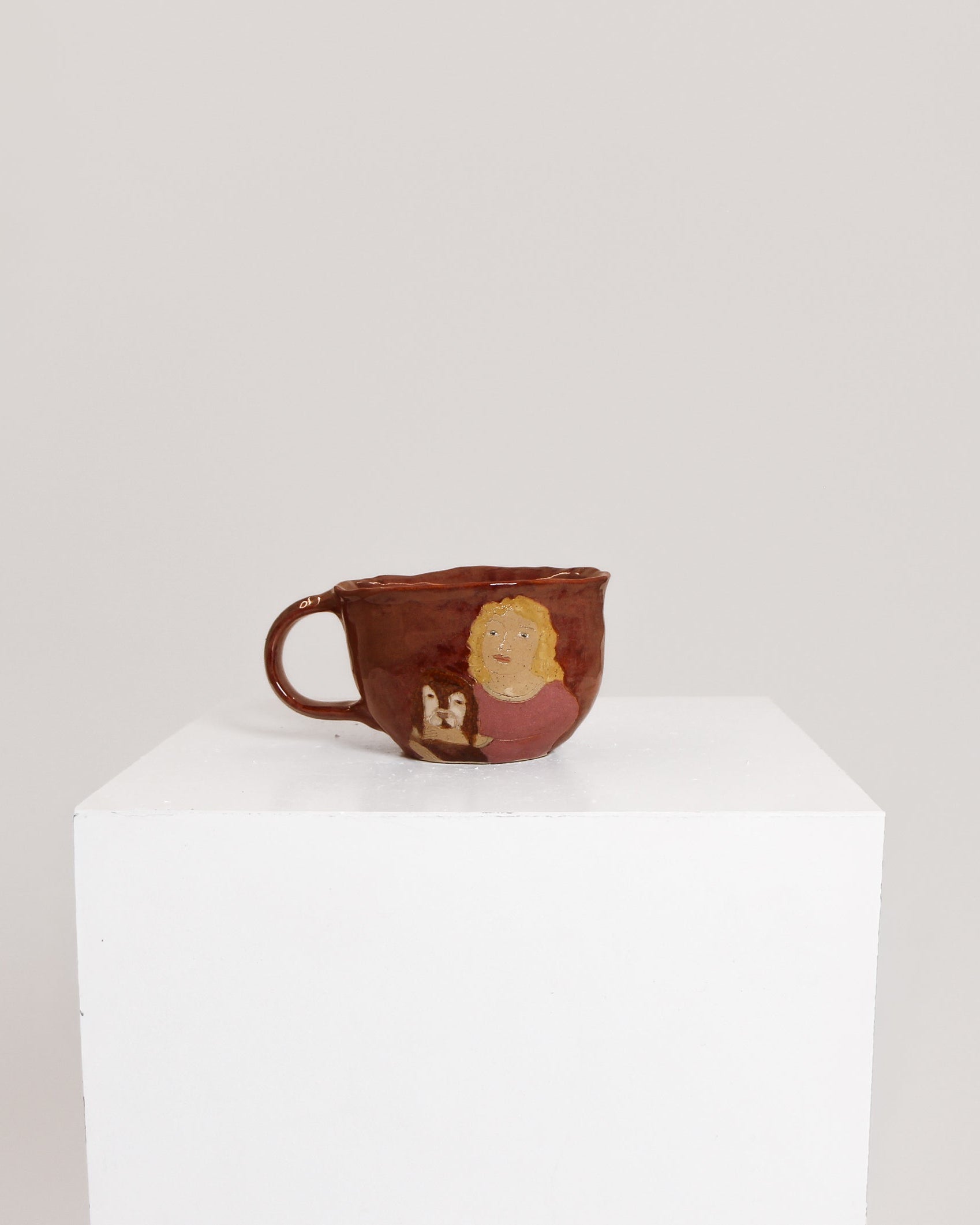 A small handmade ceramic cup with a handle, featuring a rustic reddish-brown glaze and a hand-painted illustration of a face and figures, displayed on a white pedestal against a light gray background.