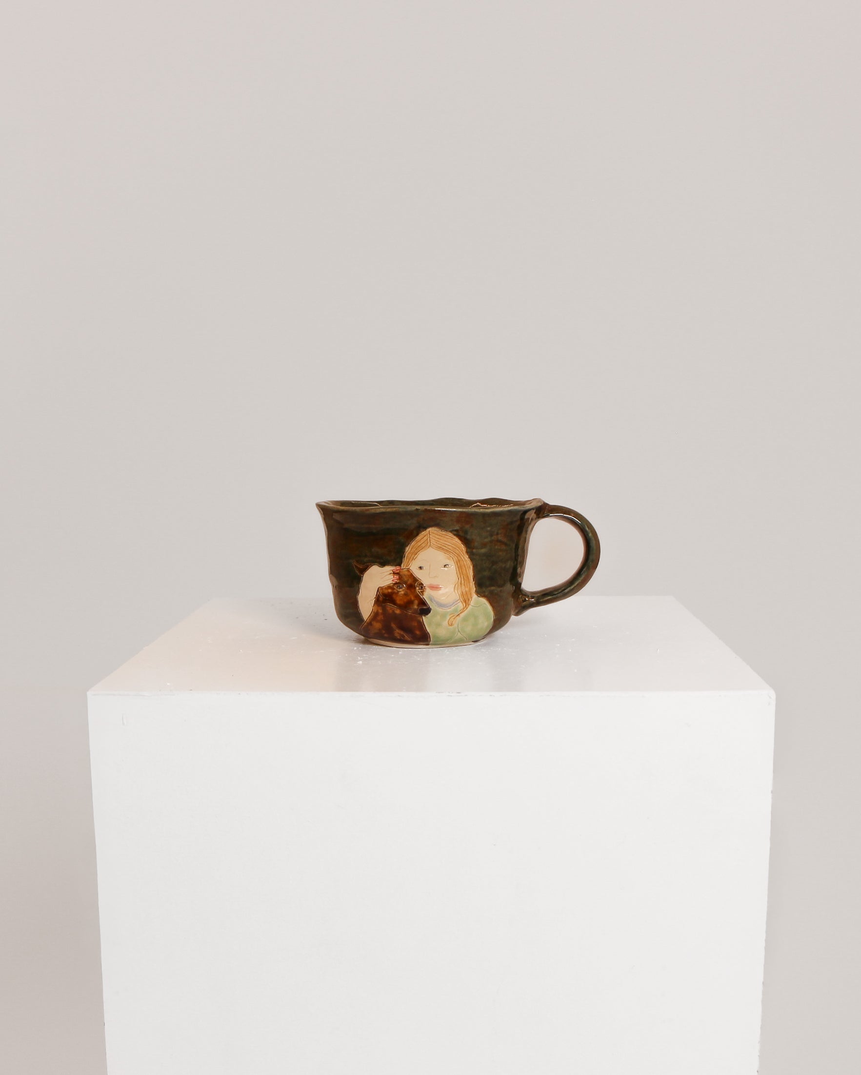 A small dark brown ceramic cup with a painted illustration of a woman and child, displayed on a white cube pedestal against a light gray background.