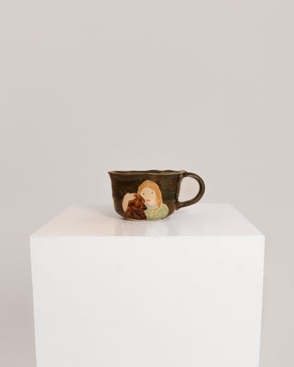 A small dark brown ceramic cup with a painted illustration of a woman and child, displayed on a white cube pedestal against a light gray background.
