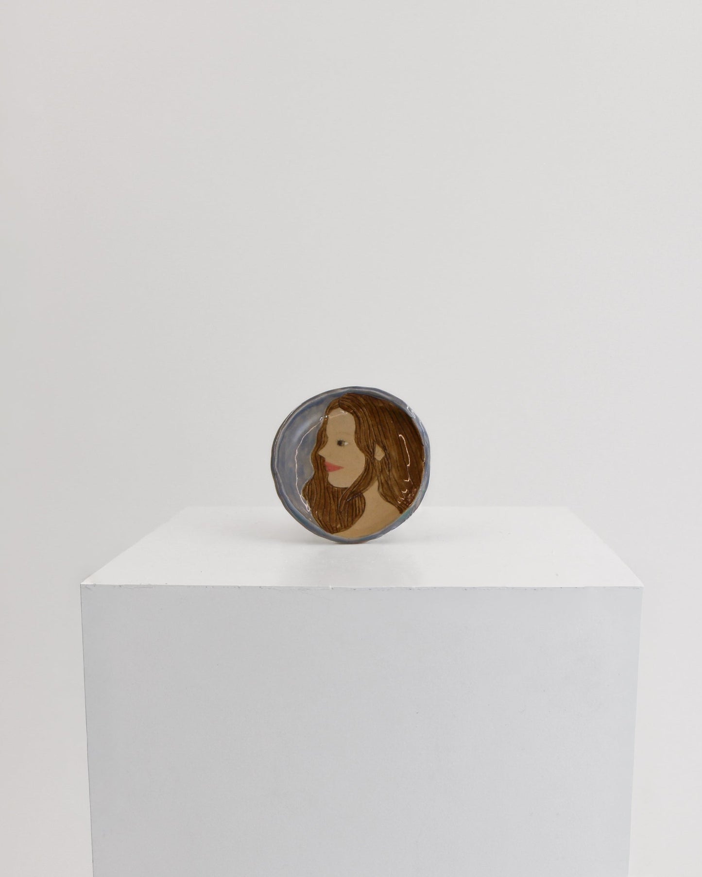 A small circular product with a portrait illustration of a woman, displayed on top of a white rectangular pedestal against a plain light gray background.