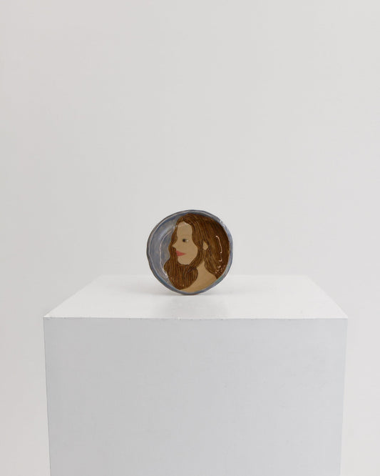 A small circular product with a portrait illustration of a woman, displayed on top of a white rectangular pedestal against a plain light gray background.