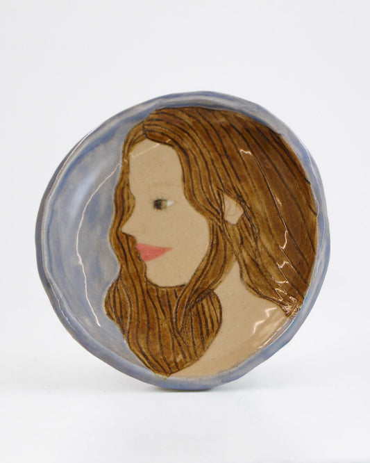 A round handmade ceramic plate featuring a painted portrait of a woman with long brown hair, set against a plain white background.