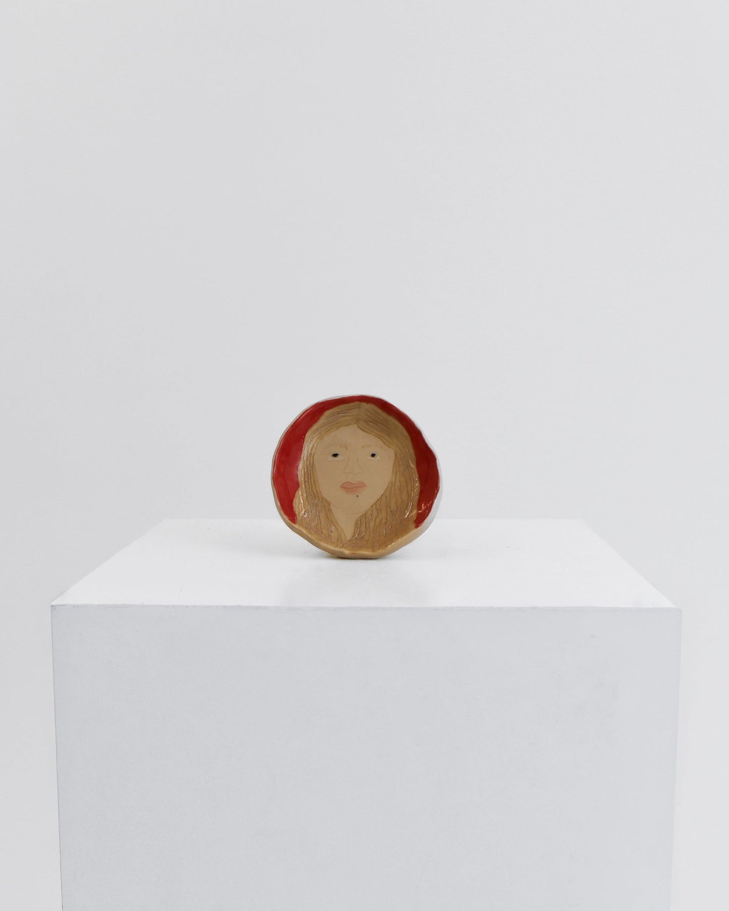 A small round wooden figurine depicting a stylized face with long light brown hair, set against a solid red background, displayed on a white cube pedestal.