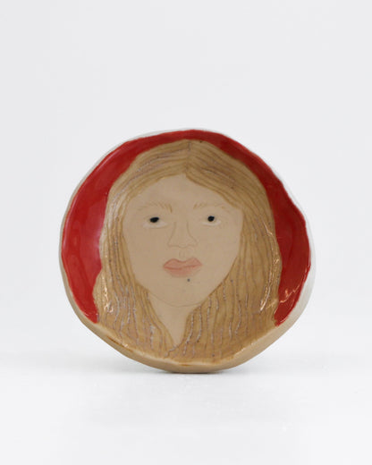 A round ceramic plate featuring a stylized portrait of a woman with blonde hair, black eyes, and pink lips, set against a red background, photographed on a white studio background.