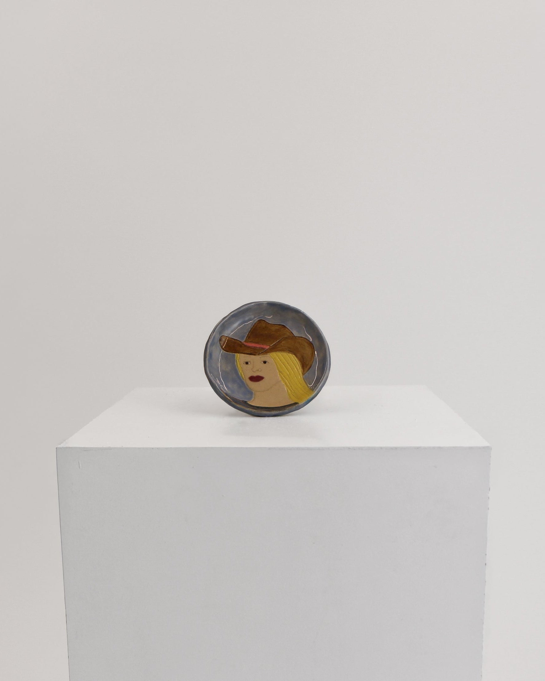 A small oval dish featuring a stylized face with a brown hat, displayed on top of a white rectangular pedestal against a light gray background.