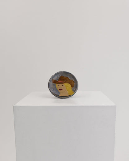 A small oval dish featuring a stylized face with a brown hat, displayed on top of a white rectangular pedestal against a light gray background.