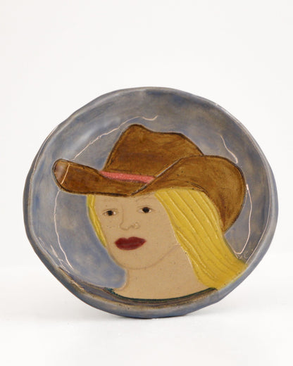 A round ceramic plate featuring a painted portrait of a woman with blonde hair, a brown cowboy hat, and red lips, set against a plain white background.
