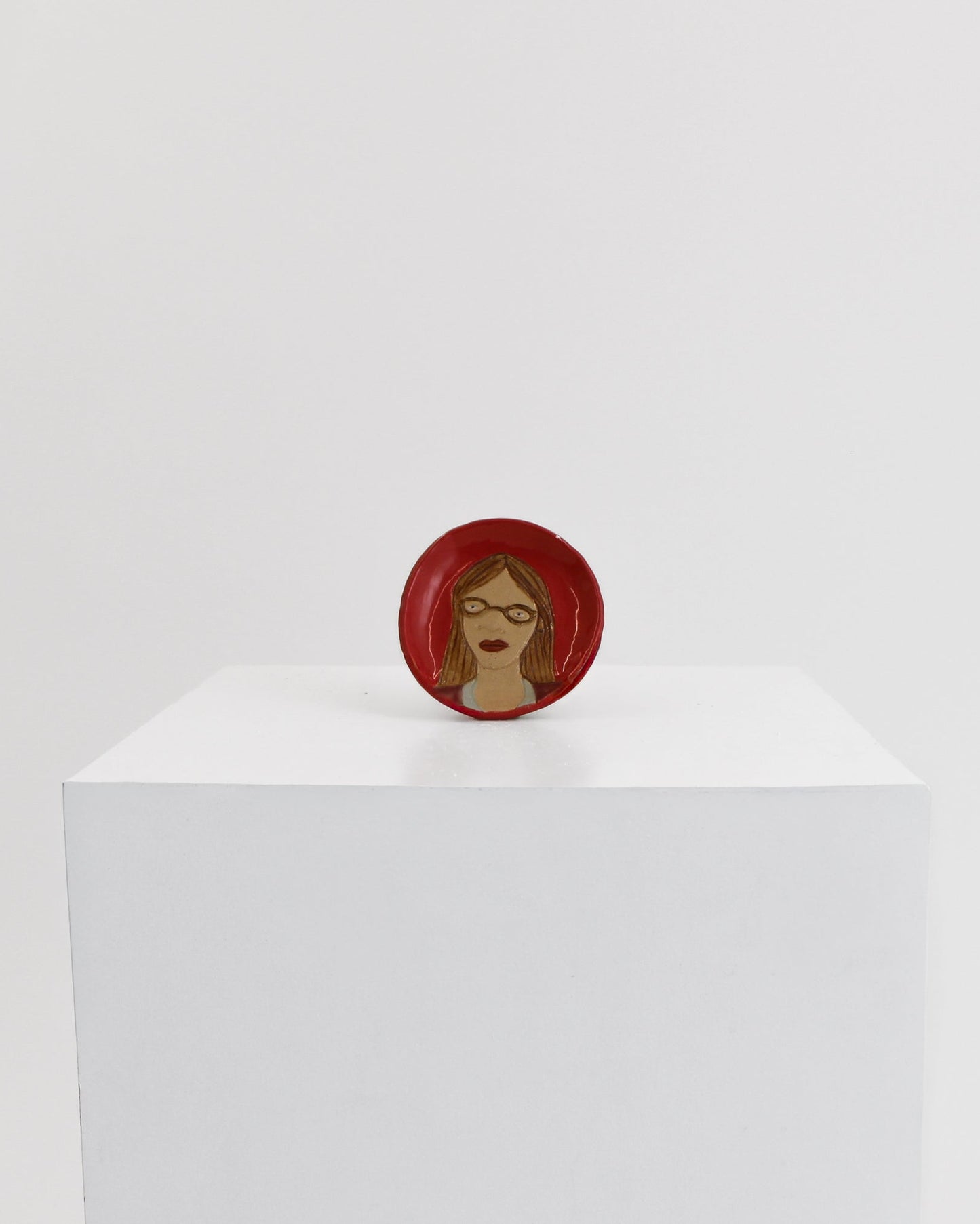 A small circular red pin or badge featuring a stylized face with brown skin and red hair, displayed on top of a white cube against a light gray background.