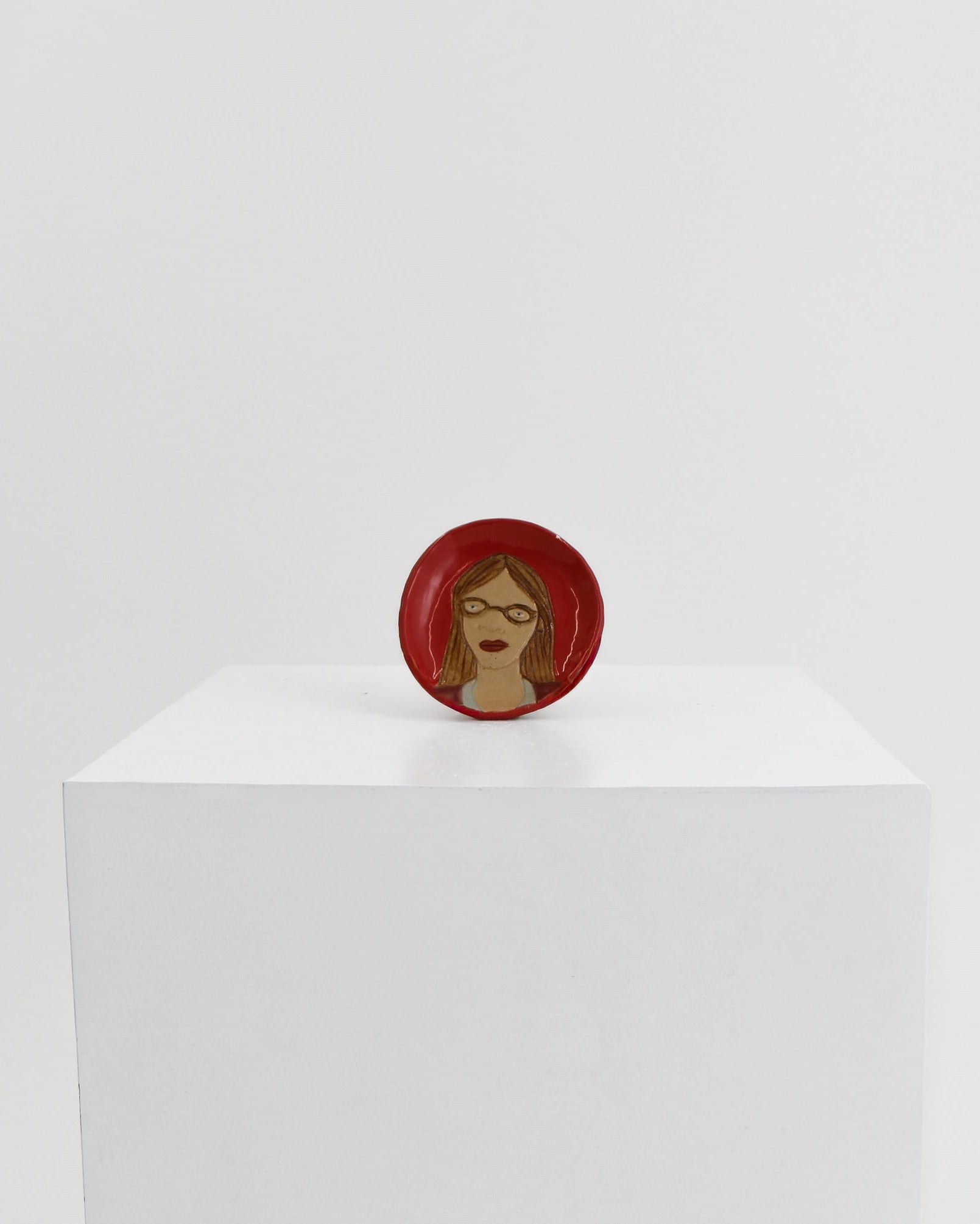 A small circular red pin or badge featuring a stylized face with brown skin and red hair, displayed on top of a white cube against a light gray background.