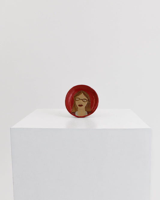 A small circular red pin or badge featuring a stylized face with brown skin and red hair, displayed on top of a white cube against a light gray background.