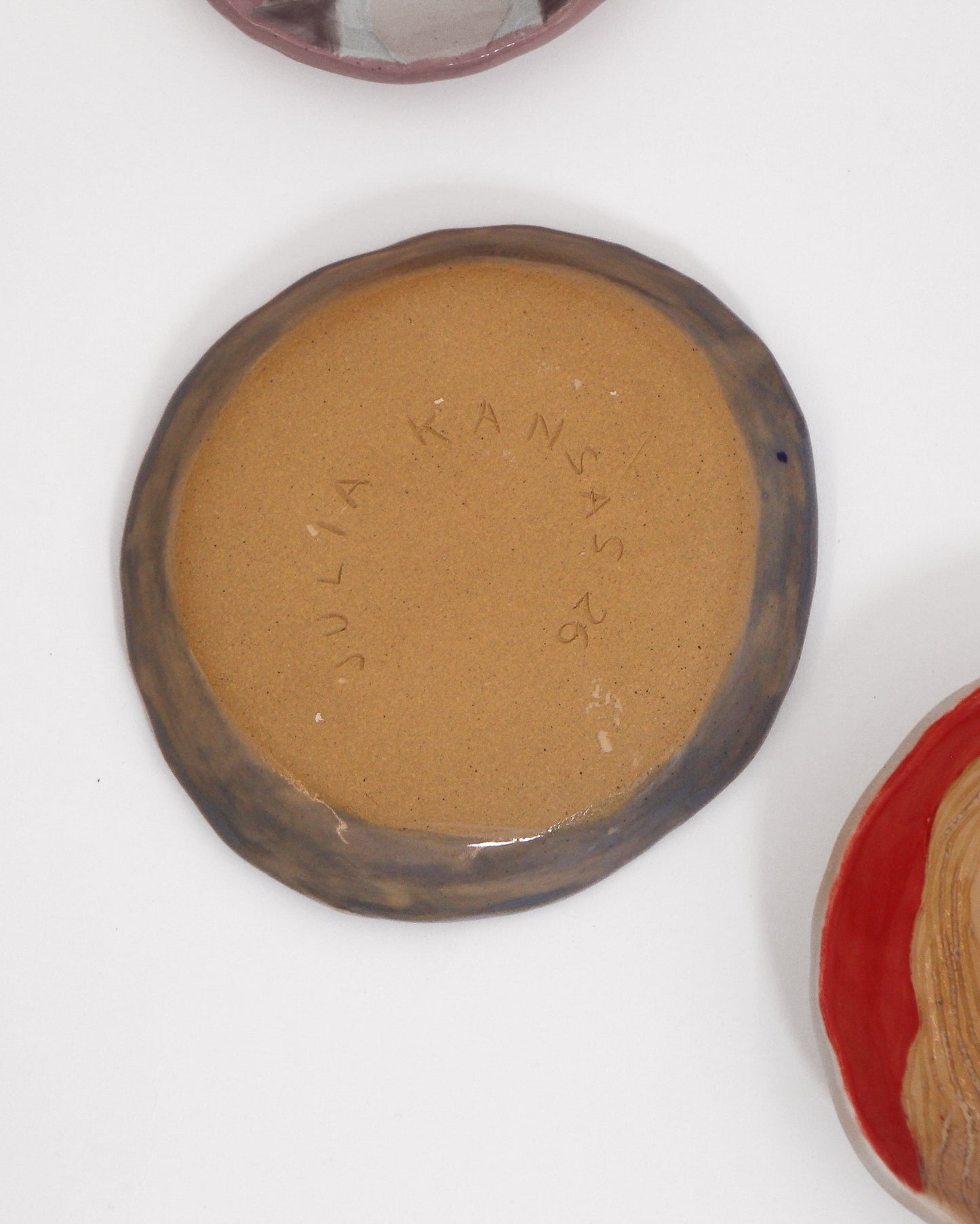 A handmade ceramic dish with a brown glazed rim and a tan back. Another partially visible ceramic tin with a portrait illustration is on the right, all set against a plain white background.