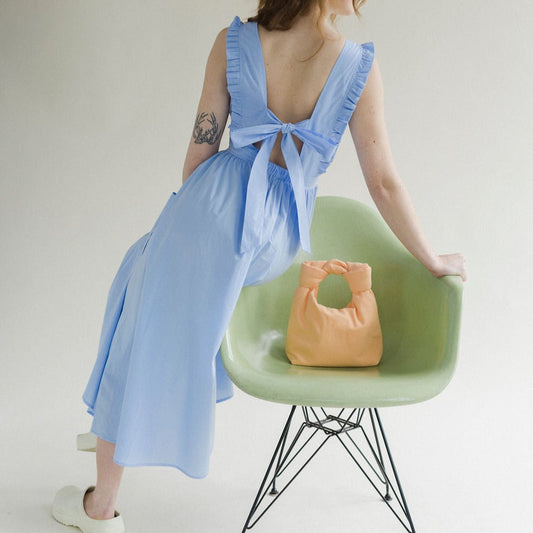 Light blue coloured apron dress with pockets, defined waist band, large straps at back to tie as bow detail, ruffled shoulder straps, and square neckline