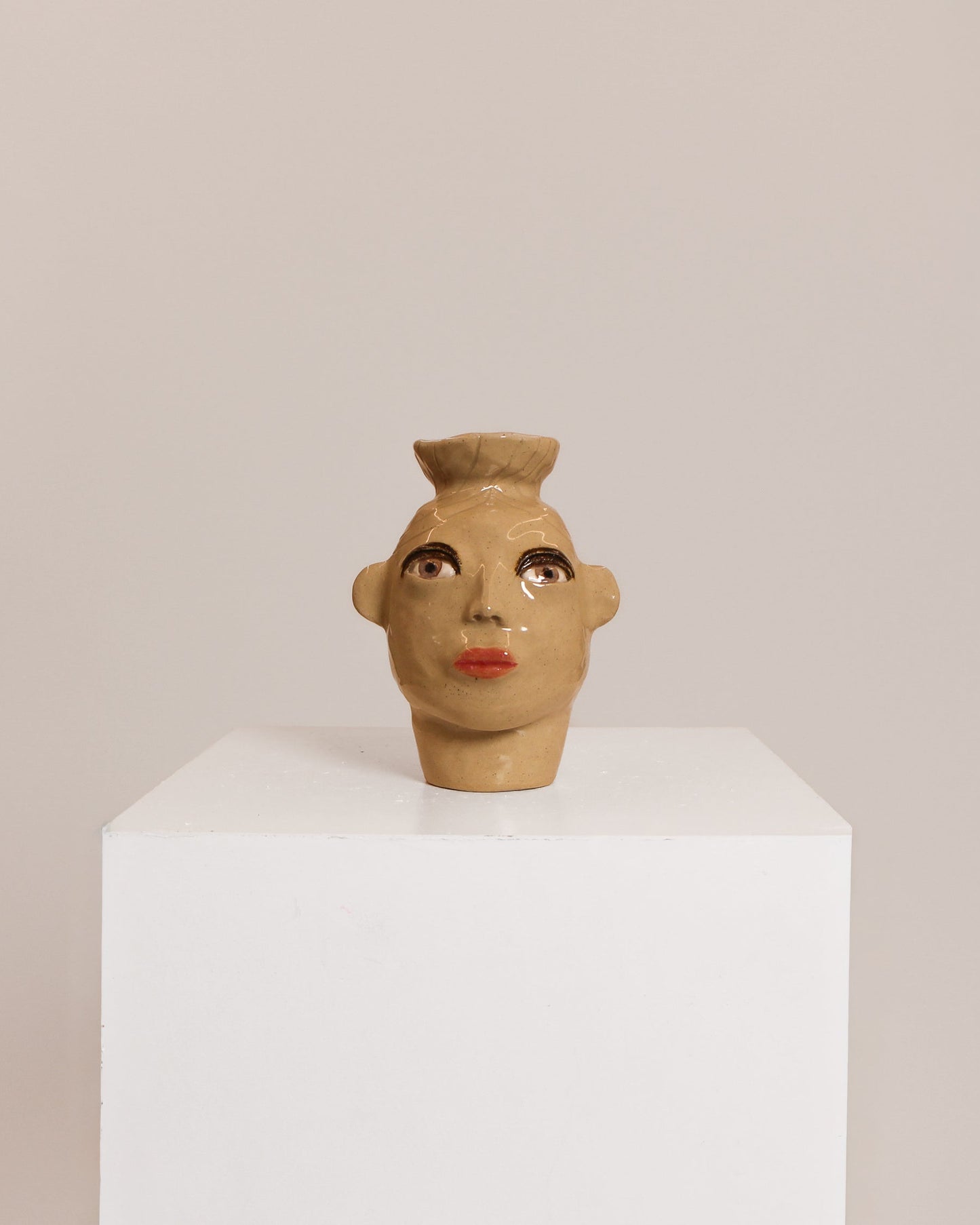A small sculptural ceramic head with a stylized face and topknot, displayed on a white pedestal against a plain beige background.